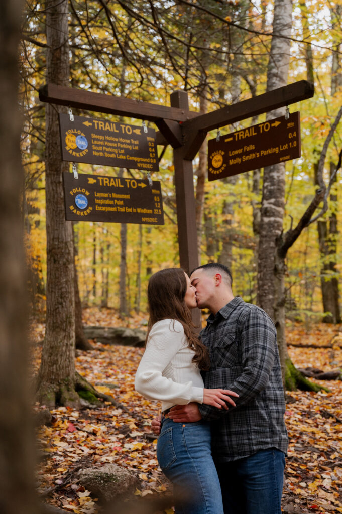 Aubrie and Dolan sharing a kiss underneath the trail signs at Kaaterskill Falls during their engagement session in the Catskill Mountains.