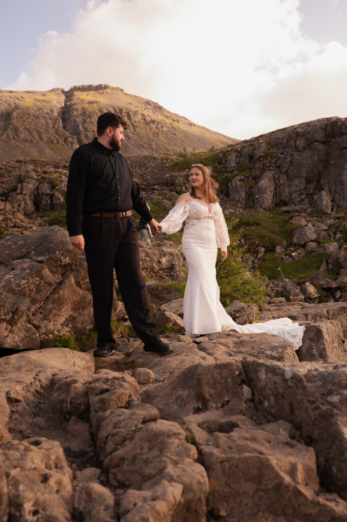 The bride and groom holding hands on a rock at Dynjandi Waterfall for their Iceland destination wedding photos.